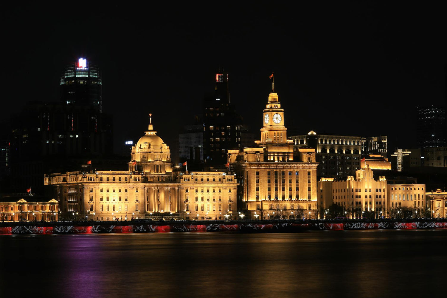 Illuminated historic buildings along The Bund in Shanghai at night, reflecting over water.